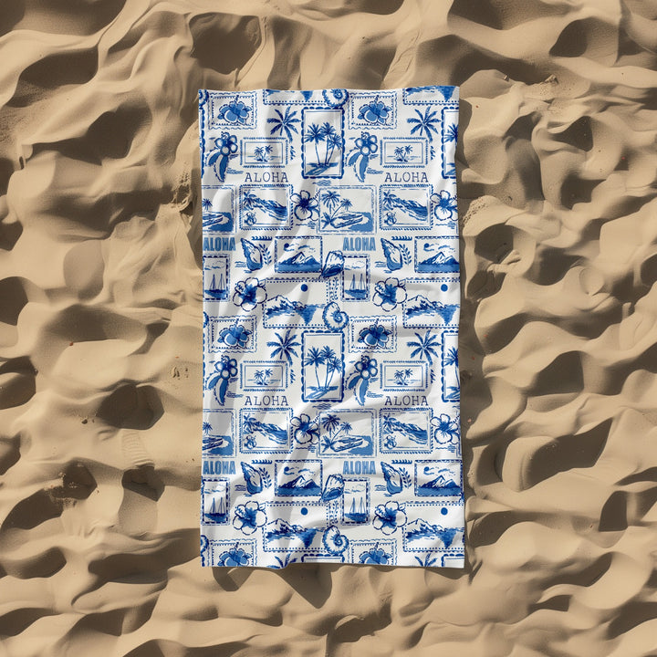 Blue and white patterned towel with 'Aloha' text on a sandy background